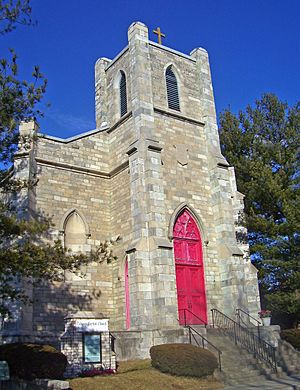 A narrow light brown stone building with a red door in a pointed arch. Above it is a square tower with a cross on top. A small sign in front says "Calvary Baptist Church".