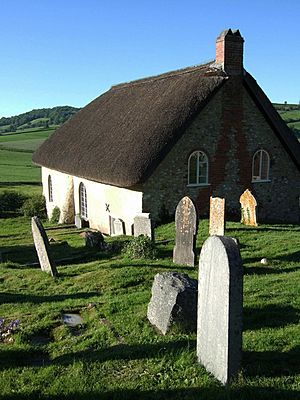 A large thatched building surrounded by gravestones, set into a hillside which slopes down towards green fields