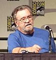 Head and upper torso view of a lightly-bearded, middle-aged man in glasses and a plain T-shirt, sitting behind a table with a microphone on it.  Two small posters that read "Comic Con International" hang on the wall behind.