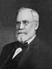 Head and shoulders of an older white man with full beard and wire-framed glasses, wearing a dark suit and bow tie.