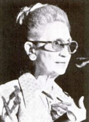An older woman in harsh light, grey hair in a bouffant updo, wearing tinted glasses and speaking at a microphone