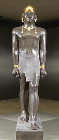 Statue of Taharqa. His name appears on the center of his belt: 𓇿𓉔𓃭𓈎 (tꜣ-h-rw-q, "Taharqa"). The statue is 2.7 meters tall. Taharqa has a striding pose, the arms held tight, and holds the mekes staff. He wears a shendyt or pleated kilt and on his head is a double-uraeus skullcap, possibly signifying his rule over Nubia and Egypt. (Louvre Museum, color reconstruction of the jewelry through pigment analysis).