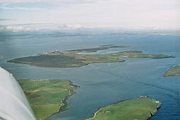 An aerial view of Flotta from the south-west. The oil terminal is visible to the centre-left, with the airport further to the left. South Walls is at the bottom and bottom left, and Switha at the extreme right.