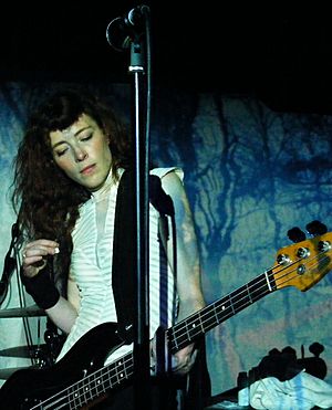A female musician performing with a bass guitar against a black and blue backdrop. A microphone is visible above her.
