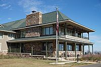 Wooden two-story lodge with green roof, large field stone fireplace, double-decker porch facing a mountain vista.