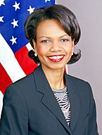 Condoleezza Rice smiling wearing a dark blue jacket over a patterned blouse. The United States flag is in the background.