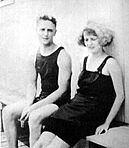 A black and white photograph of Scott and Zelda Fitzgerald in bathing suits. They are sitting on a wooden bench and leaning against a wall.
