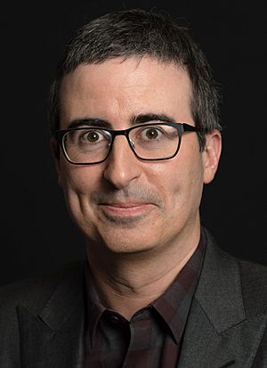 Photo of Oliver standing against a black background, wearing glasses and a dark suit jacket.