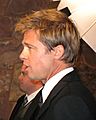 A side view of a man, who is facing to the left, with light brown hair. He is wearing a black suit and tie with a white shirt. Another male, also wearing a suit, is visible in the background. 