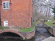 Close view of the south side of the building.  A small eliptical brick arch leads the water into the enclosed millrace, and there is an overflow weir to the right of the building. Trees are visible on the riverbank beyond.  The water is constrained in a red brick channel with a bulnose (curved) corner.  Two scroll shaped iron 'plates' are on the upper wall, terminals for tie-rods that pass through the building. This is a winter view, and the state of the trees makes this stark. There is a small and unturbulent flow of water through the overflow weir, because the level is accurately controlled by the main weir in the old lock chamber. A precarious plank bridge, with no handrails, crosses the overflow race to reach the lockside on the right.