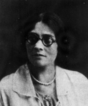 A woman with fair skin and dark hair, wearing round black glasses, pearls, and a light-colored jacket