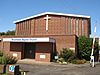 Front view of a shallow-roofed brick building with a flat-roofed entrance porch with "Broadwater Baptist Church" in blue lettering. Between this and the main roofline, there is a white crucifix, a regular pattern of single protruding bricks and a series of extremely narrow glazed ribs.