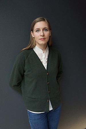 A woman with shoulder-length brown hair standing in front of a neutral background, wearing a white top, green cardigan, and blue jeans. She holds her hands together behind her back.