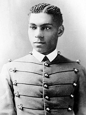 Cadet Henry O. Flipper in his West Point cadet uniform. It has three larger round brass buttons left, middle and right showing five rows. The buttons are interconnected left to right and vice versa by decorative thread. He is wearing a starched white collar and no tie. He is a lighter-colored African American with plated corn rows of neatly done hair. He is facing the camera and looking to the left of the viewer.
