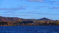 A landscape photo of Onalaska Wisconsin taken in the fall from Lake Onalaka shows a sunny sky of blue above with clouds and the cool blue waters of Lake Onalaska below in the foreground.  Sandwiched between is a horizontal ribbon that shows the city elevated 100 feet above the water and tree covered bluffs in the not to distant background.  A few houses along highway 35 are barely visible.