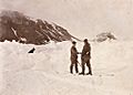 Two men shake hands in the midst of a snowfield, with a dog sitting nearby. Dark hills are shown in the background.