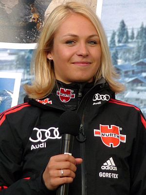 A blonde woman shown from the waist up in a predominantly black jacket, holding a black microphone, looks towards the camera and smiles slightly.