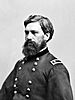 Head and shoulders of a white man with a full bushy beard and thick hair, wearing a double-breasted military jacket with a patch with two stars on the top of each shoulder.