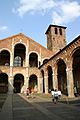 The facade and forecourt of a redbrick church are composed of simple arcades. A brick tower rises up to one side.