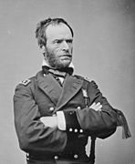 Black and white photo shows a frowning, bearded man standing with his arms folded. He wears a dark military uniform.