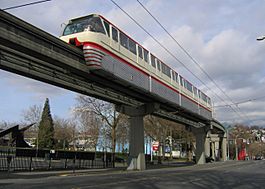 A white-and-red monorail on an elevated guideway traveling over an empty city street and parking lot