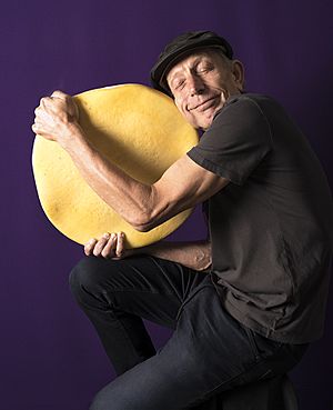 Will Studd smiling, eyes closed, wearing grey flat cap, holding a wheel of cheese about 40 cm across.