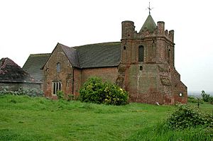 A stone church seen from the northwest, showing a squat tower with turrets on the top corners and a pyramidal roof; a transept protrudes from the centre of the church and part of the listed stable is on the extreme left