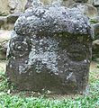 A low standing stone protruding from short grass, with the stonework of a structure visible behind it. The upper portion of the stone has a raised area perhaps representing a cap or helmet. Below this are carved two eyes in the form of circles divided by a horizontal line, giving the appearance of being closed. The cap and the area between the eyes are covered with pale lichen