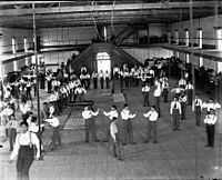 Carlisle Students in School Uniform Exercising Inside Gymnasium; Some with Indian Clubs, Others with Gymnastic Equipment; Non-Native Group Watching 1879