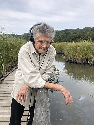 Annea Lockwood, 80 years old, stands on a boardwalk over a marsh, surrounded by reeds, and smiles as she looks at the water