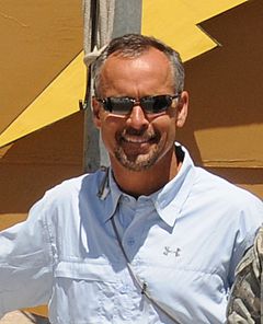 Chest-up casually posed photograph of Nolan wearing a light colored button-up shirt with an Under Armour logo and sunglasses