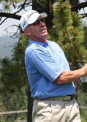 A man in a blue polo shirt and a white cap is standing on a golf course. He is wearing sunglasses as he looks after a ball he has just hit.