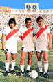 Photo of three men, wearing all-white uniforms marked by a red diagonal stripe in their jerseys, inside a stadium filled with spectators