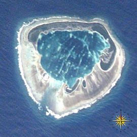 A satellite photograph of an atoll consisting of four islets with an interior lagoon. The largest island has a "C" shape and is dominated by vegetation. The other three are smaller and have sparse vegetation. The islets are surrounded by dead coral and the ocean. In the lower right corner of the photo, a compass indicates the orientation of the island.
