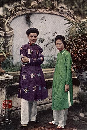Dressed in the pleated silk trousers favored by the women of Annam, a princess pauses with her husband in their tropical garden at Huế
