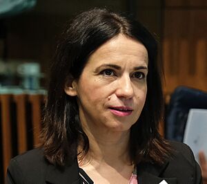 Silvia Calzón, wearing a light turquoise jacket over a black top, sits and looks slightly to the side in front of a black microphone and a pump-bottle of hand sanitizer.