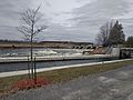 A narrow boating channel is in the foreground, past a small tree. In the background is a dam that is creating rapids in the Ontonabee River. The sky is grey, and the picture was taken in mid-April 2019.