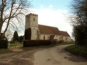 A flint church with red tiled roofs seen from the southwest, showing a tower with a battlemented parapet, the nave with a porch, and the chancel
