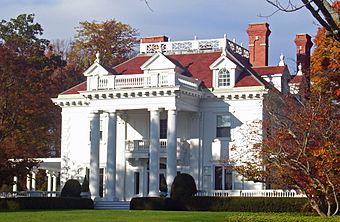 A white house with a colonnaded front and dormer windows in its red roof with brick chimneys on top seen from slightly to the right of center. The sun lights the house from the left and on either side are trees showing fall color