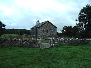 A small, simple, stone church, in front of which is a stone wall.  On the far gable is a bellcote with a single bell.