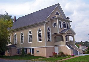 A light brown building with black roof and arched windows along the side with six-pointed stars in the tracery. Large letters on the front porch say "Agudas Achim".