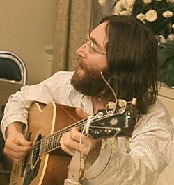 A bearded, bespectacled man in his late twenties, with long black hair and wearing a loose-fitting white shirt, sings and plays an acoustic guitar. White flowers are visible behind and to the right of him.