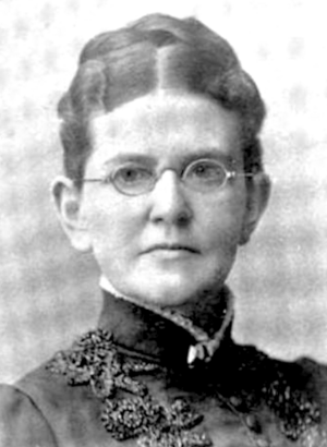 A white woman wearing glasses and a dark, high-colored dress or jacket; her dark hair is parted center and dressed in an updo