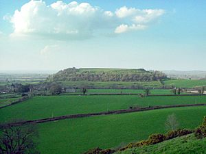 An aerial view over green fields and hedgerows toward a large conical hill