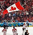 A helmetless ice hockey goaltender carrying a large Canadian flag by its pole over his head as teammates, spectators and media look on. He is wearing a white and red jersey with white pads.