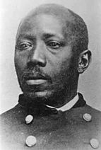 Black and white photo of a Black man of steady countenance and with short, receding hair, and a short goatee. He is wearing a military jacket over a white shirt.