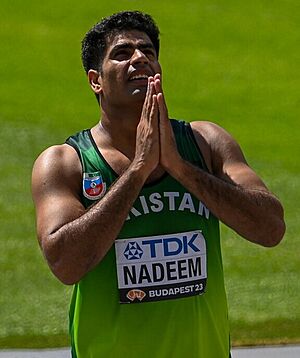 Arshad Nadeem looking up with his hands together in celebration. He is a man in his late 20s with short hair and no facial hair wearing a sports jersey.