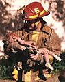 A firefighter is holding a dying toddler in his arms, and he is looking down at her. The toddler has blood on her head, arms, and legs, and is wearing white socks.