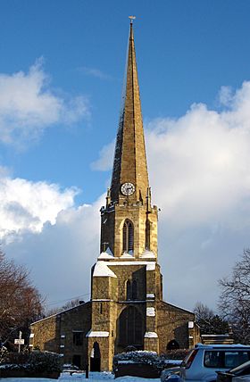 A church, about 50 metres tall, the top half of it a spire with a clock at the base of the spire. The church is lit by sunlight, with some snow on ledges around the tower, and a blue sky with a few clouds behind it