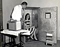 photograph of a woman, sitting on a chair which is in a large, wardrobe-sized box, the open door of which has a small window. A man stands nearby, holding a breathing apparatus.